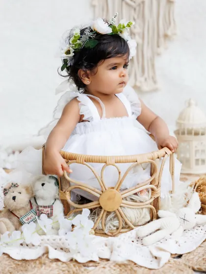 A little angel in a white dress and floral crown sits in a beautiful wicker basket, surrounded by soft, neutral props.
