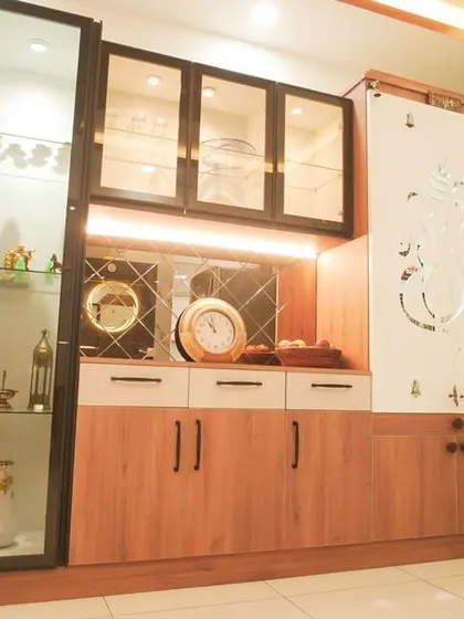 A detailed shot of the crockery and pooja unit combination. The design features a tall glass display case, under-cabinet lighting, and a beautifully carved Ganesha on the pooja door.