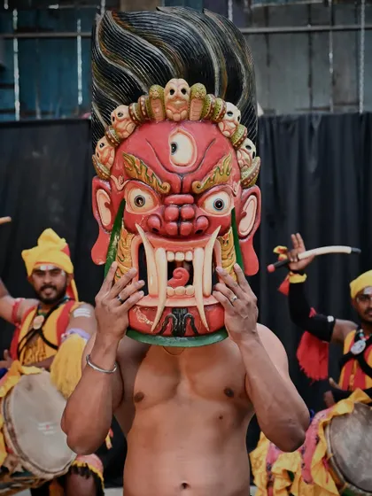 A red Nepali mask with intricate details, held by a Dollu Kunitha dancer. The vibrant colors and fierce design are meant to reflect the spirituality and heritage of Nepal, offering protection from bad luck.