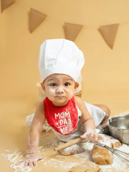 This little baker, with a dusting of flour on his nose and a cute "Mom + Dad = Me" bib, crawls towards the camera, ready for his next kitchen adventure.