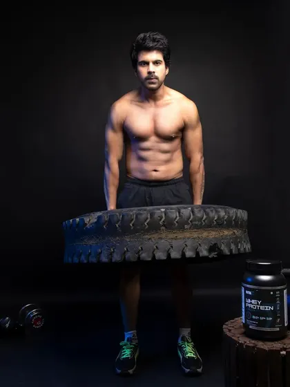 A male model demonstrating raw strength by lifting a large tire, with the whey protein product placed on a wooden stool to complete the scene.