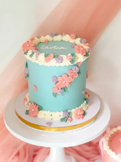 A beautiful vintage-inspired floral cake in a soft blue. The cake is adorned with a wreath of hand-piped flowers in pink, purple, and white.