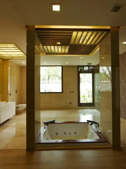 This attached bathroom in the Sachdeva Farmhouse is a spacious, modern retreat with a sunken Jacuzzi tub as its focal point. Warm stone finishes, wooden flooring, and patterned wallpaper create a refined, spa-like ambiance.