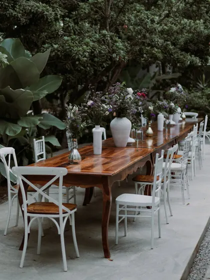 A long wooden table set for an intimate dinner, adorned with simple floral arrangements in white vases, creating a rustic and elegant look.