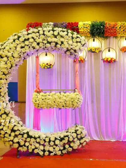 A stunning cradle ceremony setup featuring a crescent-shaped arch made of white flowers. The hanging cradle, also adorned with flowers, creates a beautiful focal point, while colorful lanterns add a festive touch.