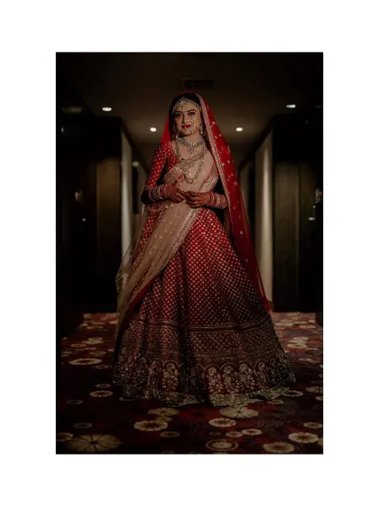 A full-length portrait of a bride in a stunning red lehenga, standing in a richly decorated hotel corridor. The dramatic lighting and composition give this a very regal and editorial feel.
