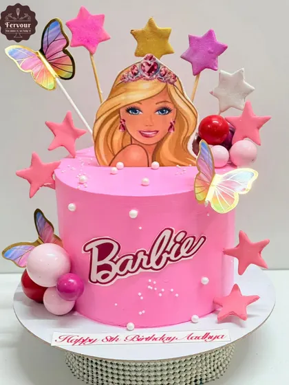 A pretty pink Barbie-themed cake decorated with stars, butterflies, and decorative spheres. The cake features a Barbie logo and a topper of the iconic doll.