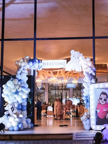 A grand entrance for a teddy bear themed first birthday. The doorway is framed by a blue and white balloon arch, with large, illuminated photo frames on either side displaying pictures of the birthday boy.
