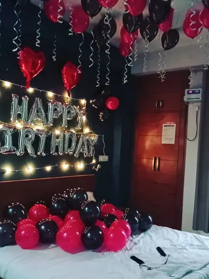 A classic birthday surprise decoration for a bedroom. This setup includes 'HAPPY BIRTHDAY' foil balloons on the wall with fairy lights, and a combination of red and black balloons on the ceiling and bed.