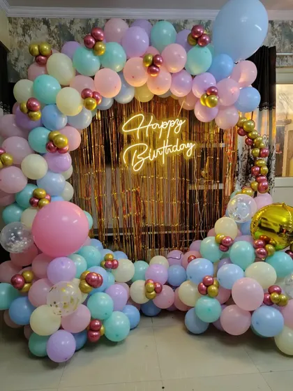 A beautiful pastel-themed birthday backdrop. The setup includes a semi-circular balloon arch over a gold shimmer curtain with a neon sign.