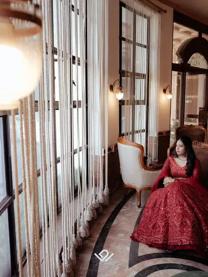 A wide shot of a bride in a red gown, seated in an elegant room with large windows.
