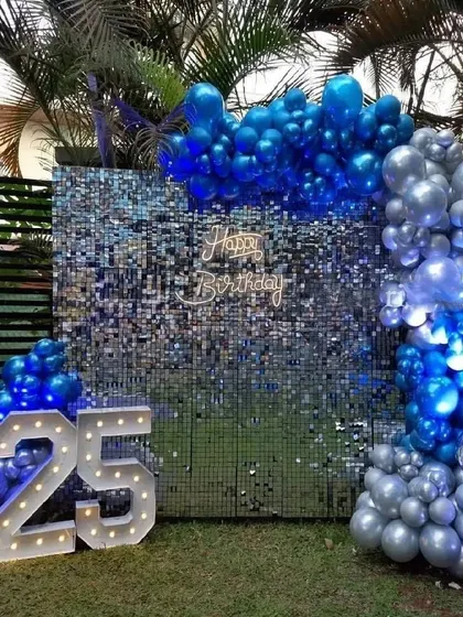 We take the party outdoors with this bold and vibrant setup on a lawn. A deep blue shimmer wall is framed by an arch of metallic blue and silver balloons, complemented by light-up marquee numbers to celebrate a milestone 25th birthday.