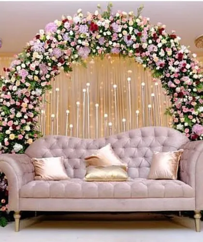 A romantic reception stage with a large floral arch. The pastel pink sofa and soft, candle-like lighting create a dreamy and intimate setting.