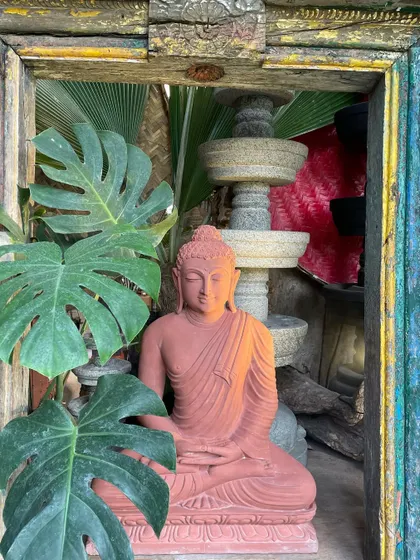 A serene Buddha in meditation, carved from red sandstone and framed by a vintage door and monstera leaves. This composition of art, architecture, and nature creates a space of boundless light and calm.