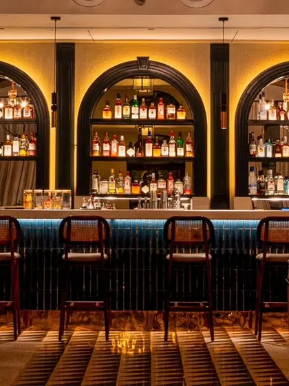 This image frames the beautifully designed bar at Vamos Experience from a distance. It shows the bar as an anchor point within the larger room, highlighting its arched shelves and warm lighting as a key feature of the restaurant's interior.