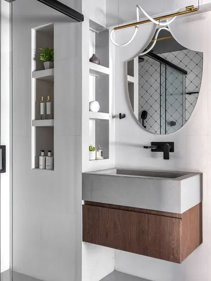 This bathroom combines a concrete-finish sink with a warm wood vanity. The unique shield-shaped mirror and modern light fixture add an artistic touch, while the built-in shelving provides practical storage.