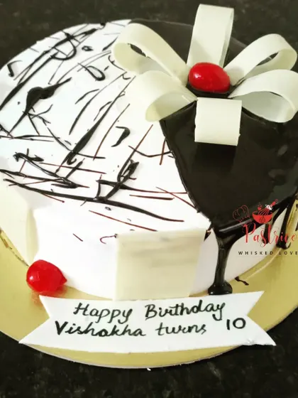 A modern black and white cake with a chocolate drip and a white chocolate bow, topped with a cherry.