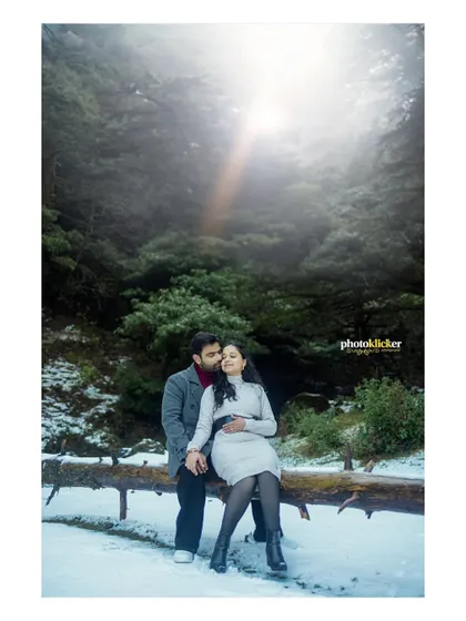 A couple shares a cozy moment sitting on a log in a snowy forest, with a soft light filtering through the trees, creating a magical winter scene.