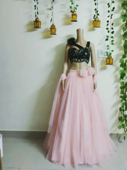 A modern lehenga design with a black embroidered one-shoulder top and a soft pink tulle skirt. The floral details on the top and waist add a fun touch.