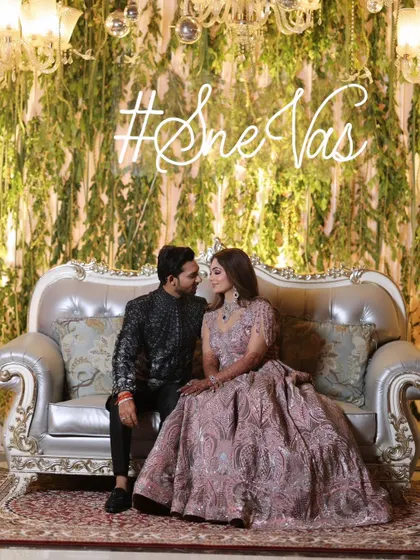 The couple seated on a silver throne-like sofa, under a neon sign of their wedding hashtag. This is a modern and stylish take on the classic stage portrait.