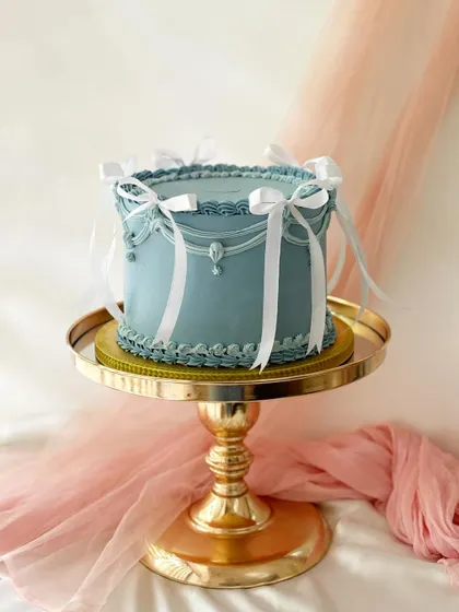 A stunning vintage-style cake in a muted blue-grey. The intricate white piping and ribbon details are showcased beautifully on a gold pedestal.