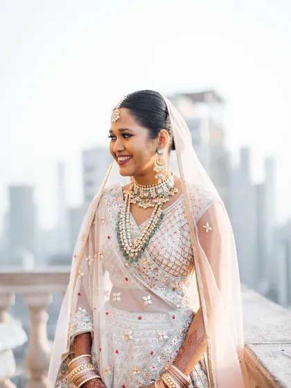 A stunning bridal portrait against a city skyline. Her hair is pulled back into a neat, low bun, allowing her sheer dupatta and beautiful jewelry to take center stage, creating a look of pure grace.