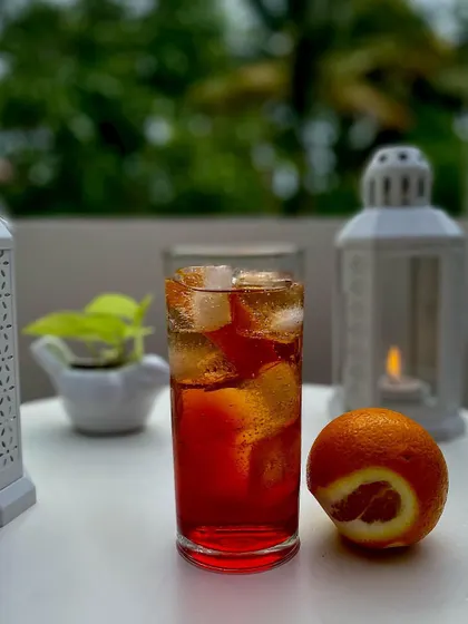 An elegant, deep red cocktail served in a highball glass with ice and an orange twist. This drink is perfect for an evening on the patio, showcasing a simple yet beautiful presentation.