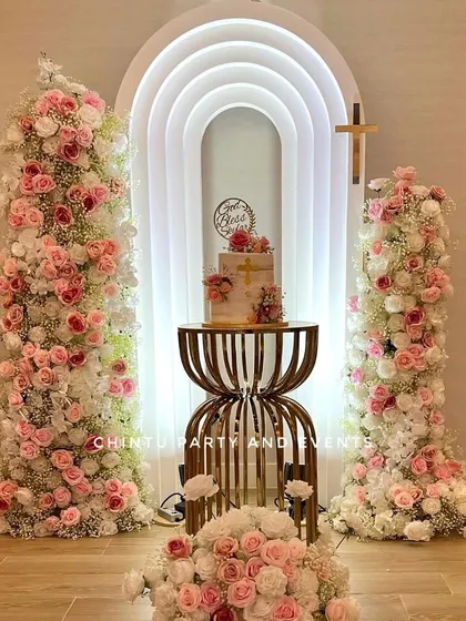 An elegant christening decoration for a girl. The centerpiece is a modern, lighted rainbow arch flanked by tall towers of pink and white flowers.