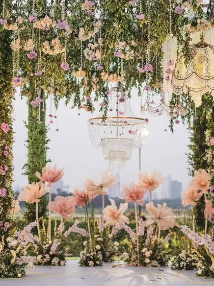 A frontal view of the open-air mandap, showcasing the harmony between the lush floral pillars, the whimsical oversized paper flowers, and the urban cityscape in the distance. This design blends nature with modern romance.