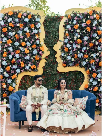 The couple poses for a formal portrait on a blue sofa against a stunning floral wall, a moment of calm amidst their vibrant Haldi celebration.