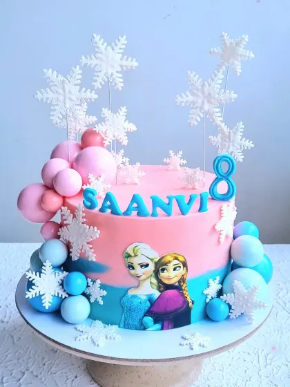 A pink and blue Frozen-themed cake decorated with edible character images, fondant snowflakes, and balloon-like spheres.