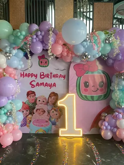 A Cocomelon first birthday party setup with a pastel color palette. The decor includes a custom backdrop with characters, a light-up number '1', and a full balloon arch in soft shades of purple, pink, and mint green.