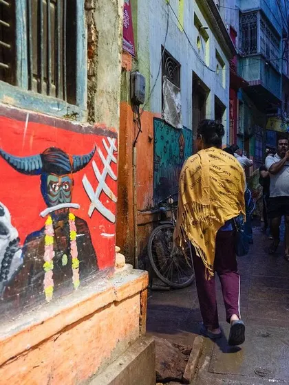 A funeral procession moves through a narrow alley in Varanasi, passing by a vibrant wall mural depicting Hindu deities. This photograph captures the journey to the ghat and the colorful context of the city's streets.
