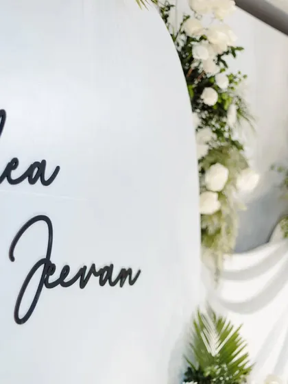 A close-up shot of the personalized backdrop for Rhea and Jeevan's engagement. The clean, modern font for their names adds a stylish touch to the white and green floral decor.