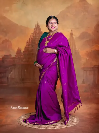 A full-length portrait showcasing the elegance of a traditional maternity look. The rich purple saree stands out beautifully against the artistic studio backdrop.