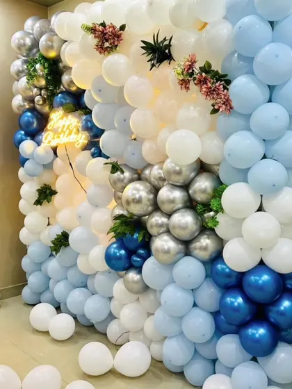 A stunning floor-to-ceiling balloon wall in shades of blue, white, and silver. A "Happy Birthday" neon sign is nestled within the balloons for a cool effect.