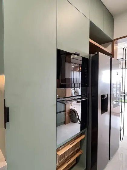 A parallel kitchen in a soothing sage green. The design includes a tall pantry unit, a roll-up shutter for appliances, and a smart refrigerator with an integrated display shelf beside it.