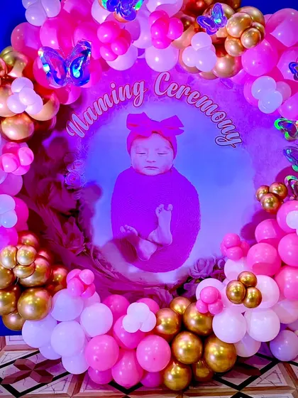 A close-up of the personalized Naming Ceremony backdrop. The baby's photo is surrounded by a dense ring of pink, white, and gold balloons with shimmering butterfly details.
