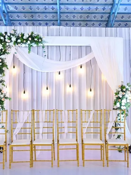 An elegant backdrop for an engagement or a small wedding gathering. The white drapes, hanging bulbs, and asymmetrical floral design create a modern and chic look.