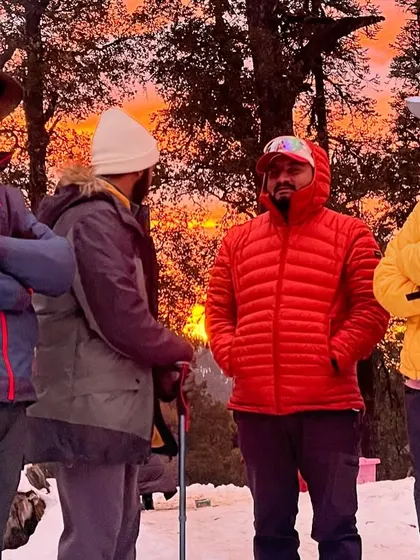 Four male trekkers in winter gear chat near a campfire against a beautiful sunset backdrop. One of the best parts of the trek is sharing stories and laughs with fellow trekkers around a warm fire after a day of hiking in the snow.