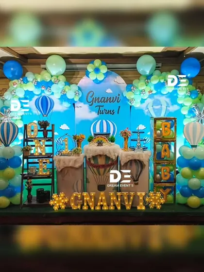 A hot air balloon themed first birthday for Gnanvi. The setup uses a green and blue color palette with balloon columns shaped like hot air balloons.