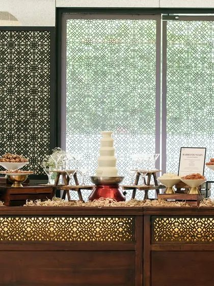 An elegant dessert buffet station with a white chocolate fountain as the centrepiece. The intricate woodwork of the counter and the symmetrical arrangement create a look of refined luxury.