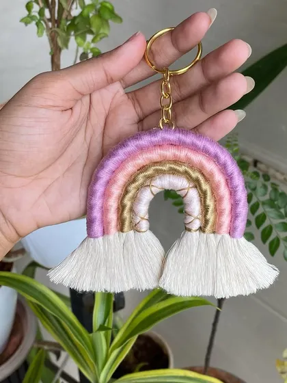 Holding up a macrame rainbow keychain in shades of pink and purple. This gives a clear view of the size and detailed wrapping of the keychain.