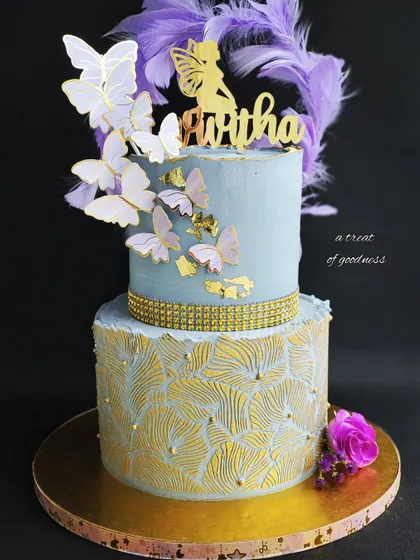 This two-tiered fairy and butterfly themed cake combines soft colors with elegant textures. The bottom tier features a gold fan-leaf stencil, while the top is a smooth canvas for a cascade of delicate butterflies.