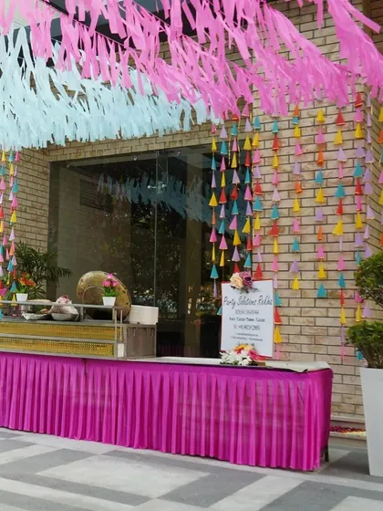 A vibrant food counter setup for a Holi party or Mehendi. The colorful hanging decorations and bright pink table skirt ensure that even the functional areas of the event are beautifully decorated.