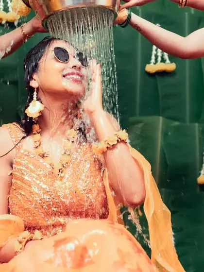 A bride enjoying her Haldi ceremony, shielding her eyes with a smile as water is poured over her.