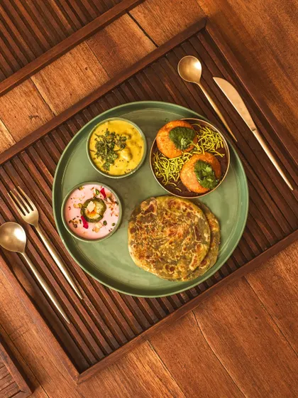 A complete meal for one, featuring parathas and various curries, elegantly arranged on a textured tray. The use of brass cutlery adds a touch of traditional luxury.