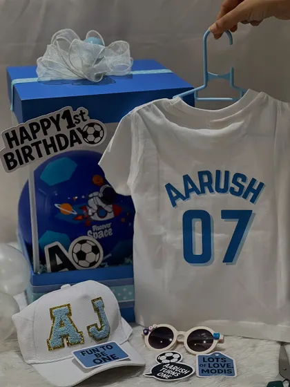 Here are all the goodies from the football-themed first birthday hamper for Aarush. It includes a personalized t-shirt, a custom cap, cool sunglasses, and a mini football, all packed in a themed box.