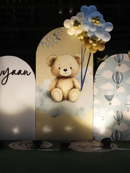 A lovely teddy bear themed backdrop for Avyaan's first birthday. The setup features three arched panels with a 3D teddy bear, hot air balloon prints, and personalized lettering.