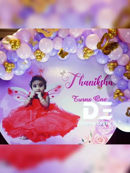 A personalized backdrop for Thaniksha's first birthday, featuring her photo in a fairy costume. The purple and gold balloon arch adds a touch of glamour.